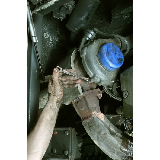 Photo of Facom 64_7_8X15_16 combination wrench used on a car exhaust manifold by a mechanic's hand.