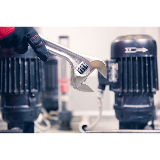 Photo of a Facom adjustable wrench 101_12 held by a gloved hand, industrial machinery background.