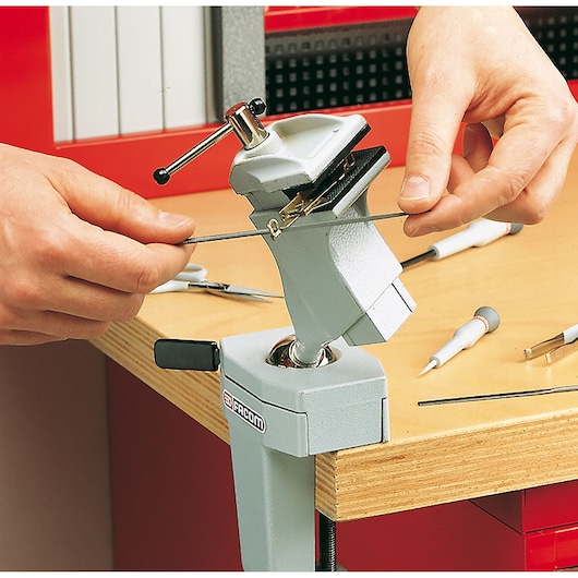 Photo of Facom 1150_MT photography bench vise clamped to a workbench, holding a file, with hands adjusting tool.