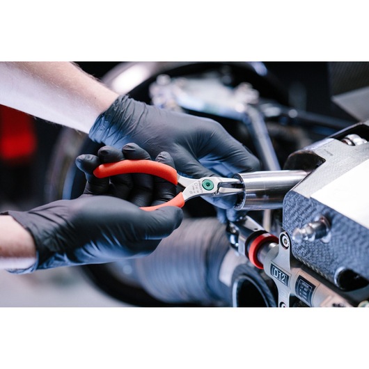 A person wearing black gloves uses red-handled Facom pliers to work on a metallic mechanical component.