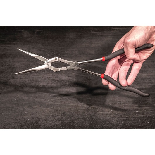 Photo of Facom 185_34L long nose pliers with black handles and red accents, held in a hand against dark background.