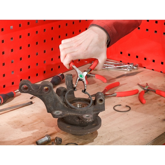 Facom 199A_23 circlip pliers with red handles used on a metal part, workshop setting with tools in background.