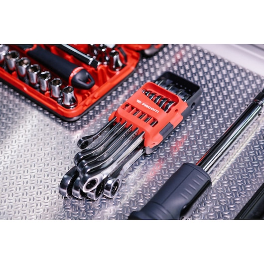 A set of Facom combination wrenches in a red and black holder, placed on a textured metal surface. Nearby is a ratchet handle and a socket set in a red case.