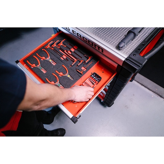 Person's arm reaching into an open drawer of a Facom tool cabinet, containing organized red-handled pliers and screwdrivers on a foam insert.