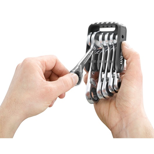 Two hands holding a set of Facom ratcheting combination wrenches organized in a black plastic holder, with one wrench being removed.