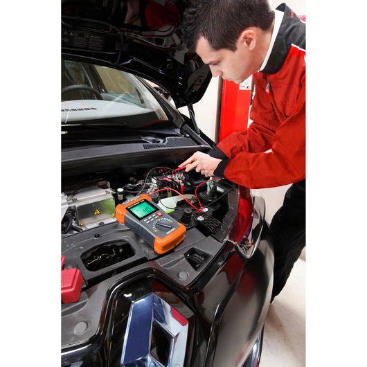 Facom 715PB automotive multimeter in use on a car engine during diagnostics, handheld device with orange accents.