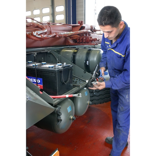 Facom 985913 tool held by mechanic in blue coveralls working on a vehicle battery and air tanks in a workshop.