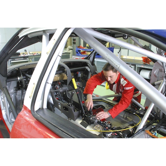 Facom A_444 technician in red suit working inside stripped race car cockpit with visible tools and roll cage.