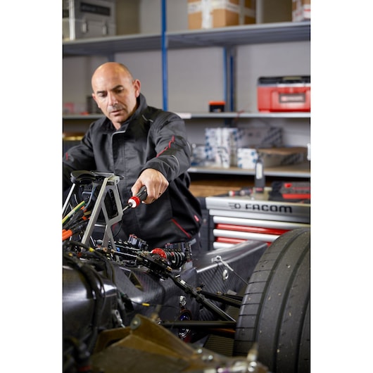 Facom AT_PA4V mechanic using hand tool on race car chassis in workshop, red Facom tool chest in background.