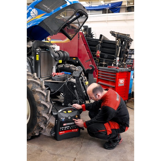 Facom B124_1A 12-24V booster in use by technician near tractor in workshop, red tool cabinet visible.