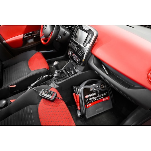 Facom 12V booster B12OBDM in car interior, placed below dashboard with cables, red and black seat and dashboard.