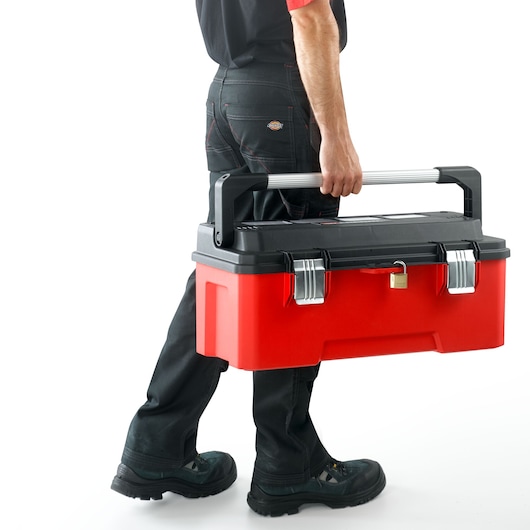 Photo of a person carrying a Facom BP.P26APB red and black toolbox with a metal handle, front view on white background.