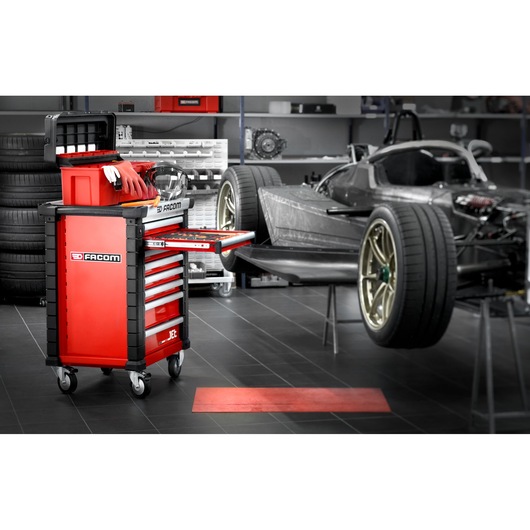 Red Facom tool chest on wheels with multiple drawers and compartments, positioned in an automotive workshop next to a racing car. The tool chest has gloves and tools visible and is organized for workshop use.