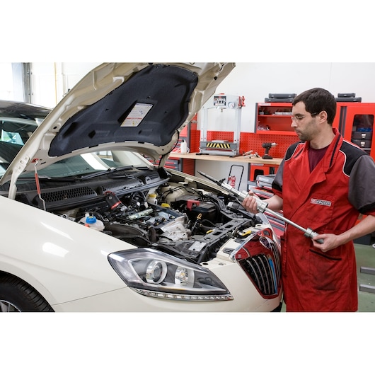 Facom DCRIP tool held by technician in red apron next to car with open hood in workshop setting.