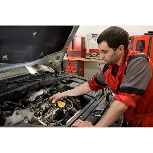 Facom DCRIP automotive technician working on car engine in workshop, red tool cabinets in background.