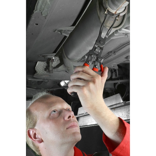 Facom DE.CPB exhaust clamp pliers with red handles used under a vehicle by a person wearing a red uniform.