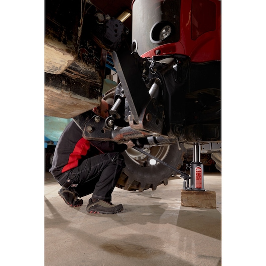 Facom DL5BTI bottle jack lifting large vehicle in workshop, worker in black and red uniform using tool, ground level view.