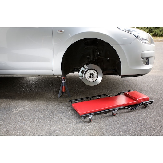 Facom DTS_1B red padded mechanic creeper with wheels next to raised car on jack stand, front angle.