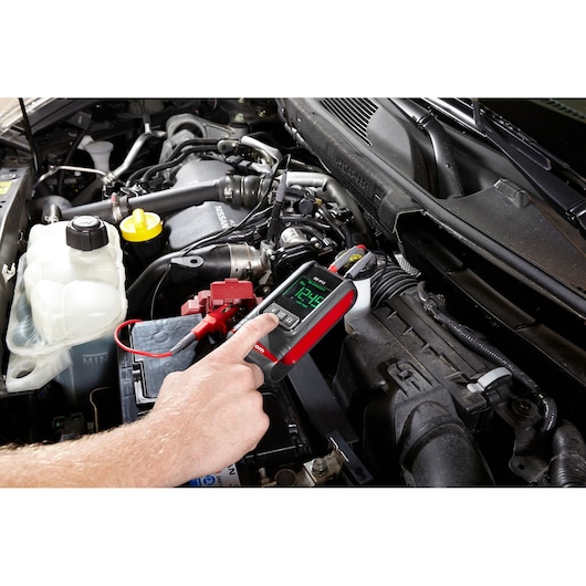 Facom DX.V12PB automotive diagnostic tool testing a car battery in engine bay, close-up view with hand operation.