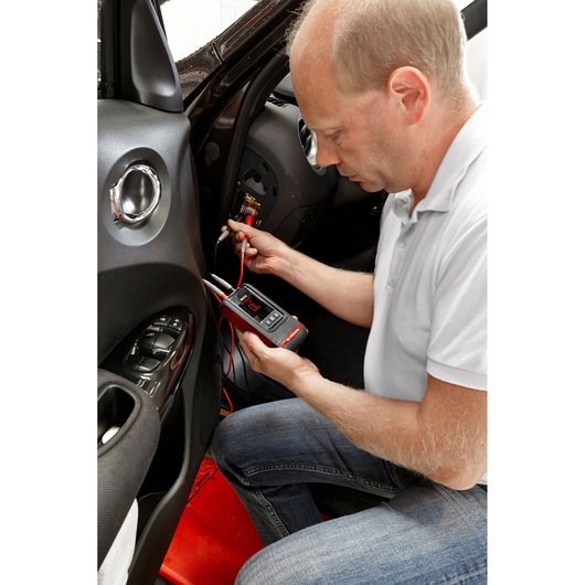 Photo of a person using a Facom DX.V12PB automotive multimeter to test a car fuse panel.