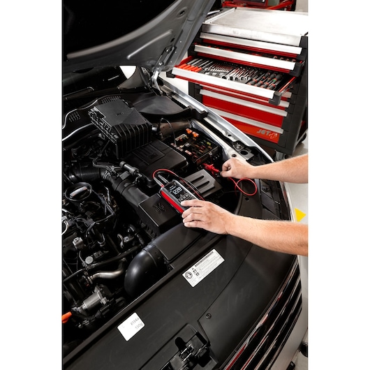 Facom DX.V12PB multimeter testing a car battery in engine bay, tool cabinet with hand tools in background.
