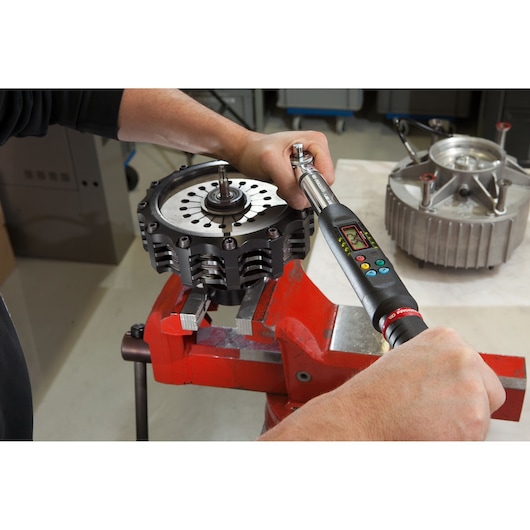 Facom E_306A135S digital torque wrench used to tighten bolts on a metal part secured in a red vise, close-up view.