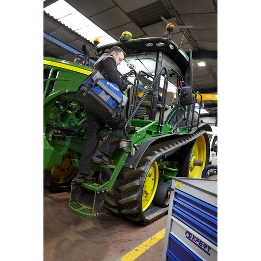 Photo of a Facom E010601 blue and black tool bag carried by a person climbing a green tractor in a workshop.
