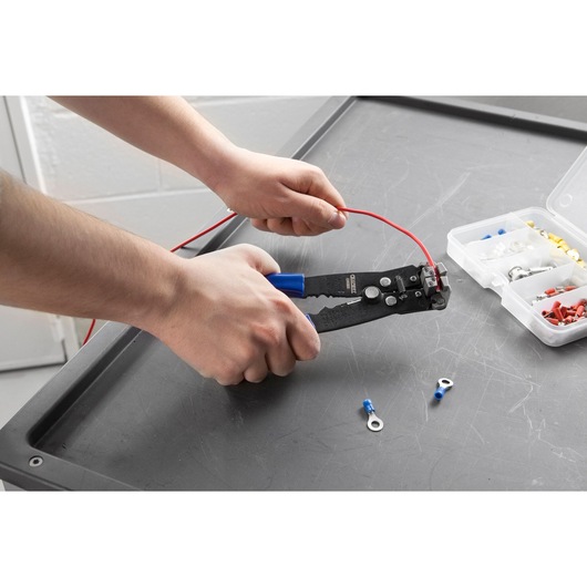Facom E050105 wire stripper with blue handles in use on red wire, box of terminals on work surface.