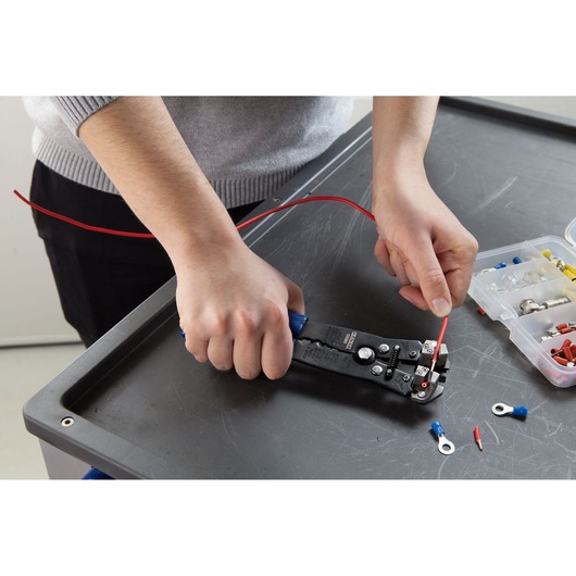 Facom E050105 wire stripper tool in use on a red wire, with electrical connectors nearby on a worktable.