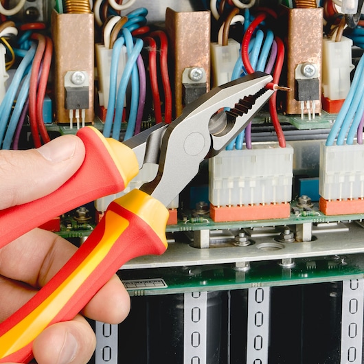 Facom E050413 insulated pliers held near electrical wires inside a circuit board, close-up.