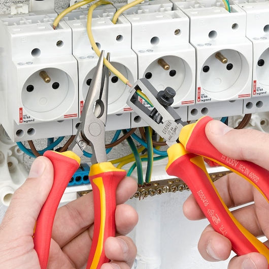 Facom E050416 insulated wire stripper and pliers held by hands near electrical outlets, front angle.