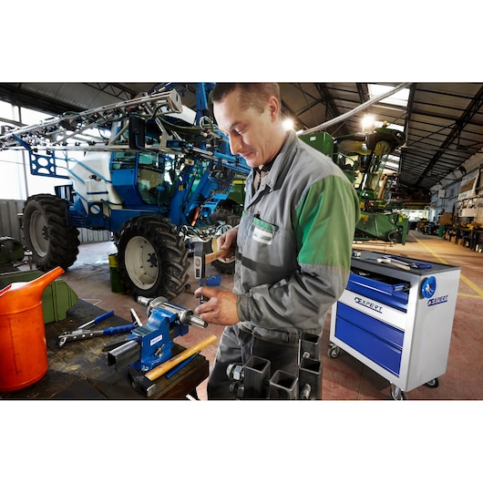 Facom E090402 workshop scene with mechanic using tools near blue rolling tool cabinet and large agricultural machinery.