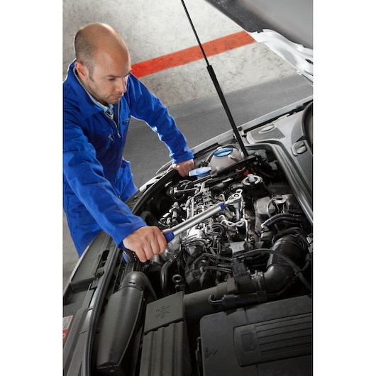 Facom E100107 torque wrench used by mechanic in blue coveralls working on car engine.