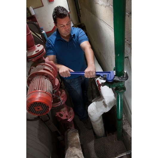 Facom E117825 pipe wrench in use on green industrial pipe, worker in blue shirt, indoor setting.