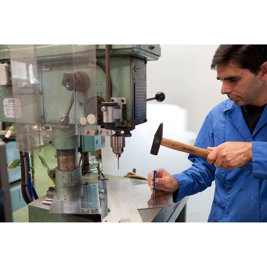 Facom E150502 machinist marking metal sheet with hammer and punch near industrial drill press.