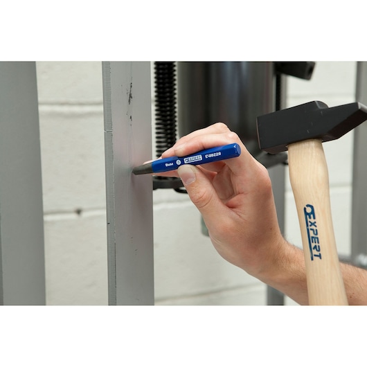 Facom E154674 metal punch tool held by hand with hammer, blue handle, workshop background.