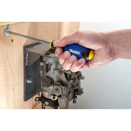 Facom E165209 hand holding blue and yellow screwdriver fastening screw into wood panel near metal machinery.