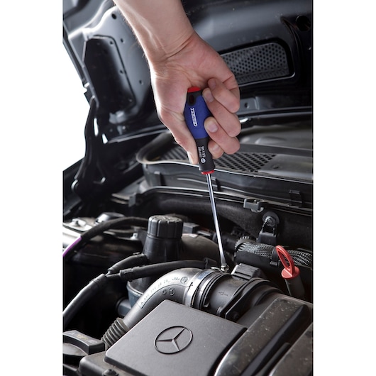 Facom E165482 screwdriver used on car engine, blue and red handle, close-up in automotive context.