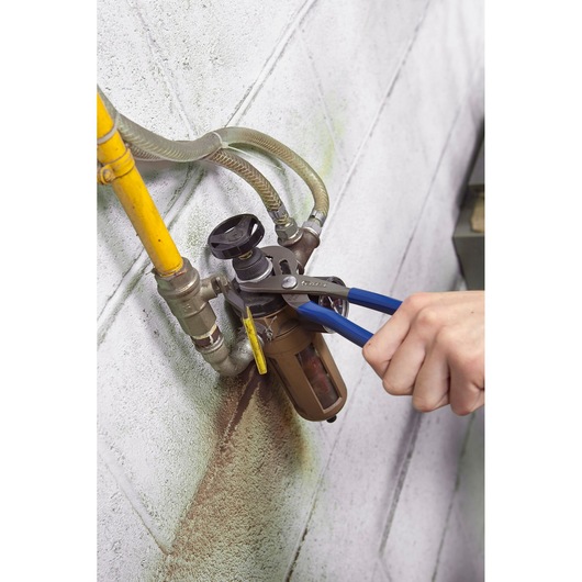 Facom E184690 pliers with blue handles used to adjust a pipe fitting against a wall.