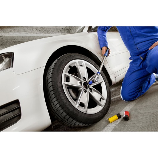 Facom E194676 torque wrench used on silver car wheel, mechanic in blue jumpsuit kneeling, tool accessories nearby.