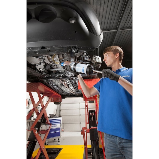 Facom E200231 mechanic using grease gun under car on lift in workshop, blue tool chest visible.