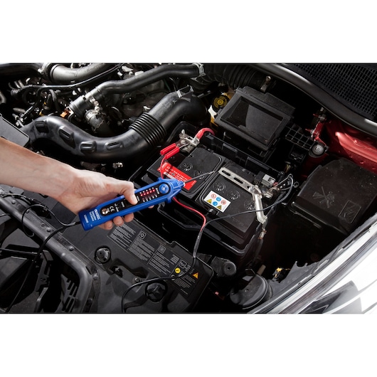 Facom E201806 voltage tester used on a car battery, hand holding tool near battery terminal.