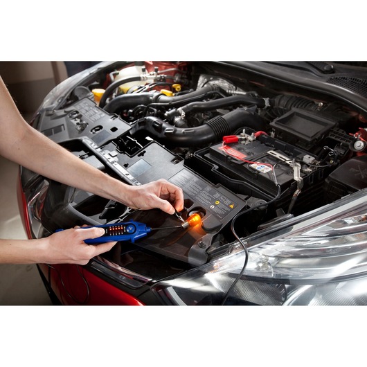 Facom E201806 voltage tester used on a car battery in an open engine bay, showing diagnostic testing.