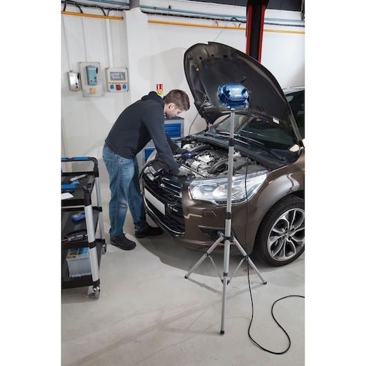 Facom E230201 portable work light on tripod illuminating car engine during repair in a garage setting, side view.