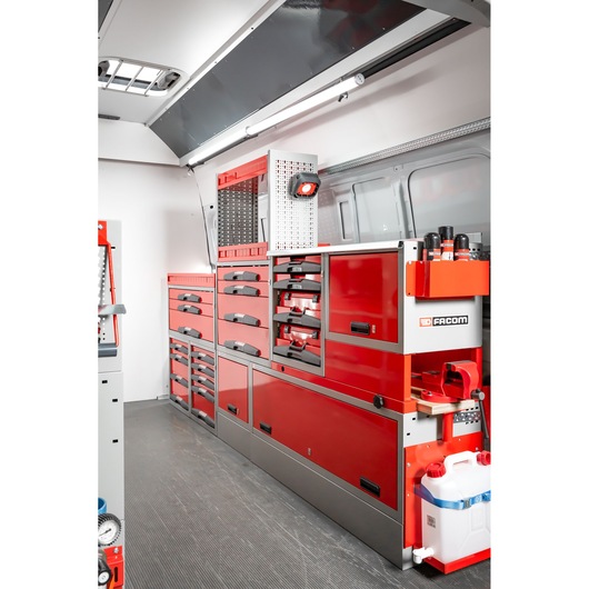 Interior view of a Facom mobile workshop setup inside a van, featuring red and gray tool cabinets, drawers, and shelving units neatly arranged along the side wall. Various tools and accessories are stored, and a white water container is visible at the bottom right.