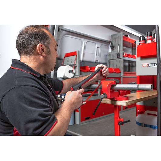 A person with a blurred face is using a hacksaw to cut a metal pipe held in a red vise inside a workshop or service van. The workspace is organized with various tools and storage units, and the Facom brand is visible on a cabinet.