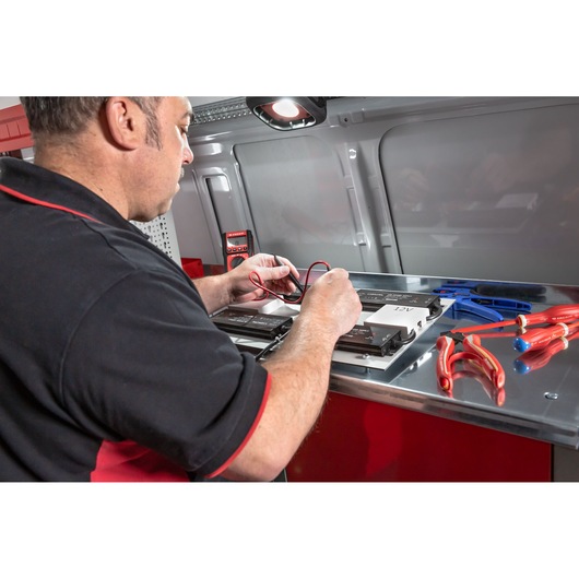 A person working with electrical tools and batteries on a metallic workbench inside a vehicle. The person's face is blurred. Various insulated red and blue hand tools and a multimeter are visible on the surface.