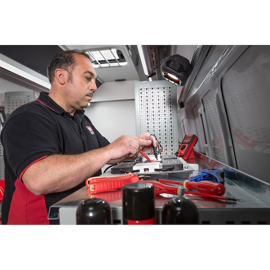 A person in a black and red shirt is using electrical testing equipment and tools inside a workshop or service van. Various hand tools, such as screwdrivers and pliers, are visible on the workbench, along with a digital multimeter.