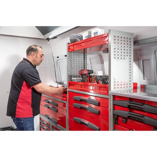 A person is working inside a vehicle equipped with red Facom storage cabinets and tool drawers. Various tools, including a drill, are visible on the shelves. The individual is wearing a black and red shirt and is accessing the drawers.