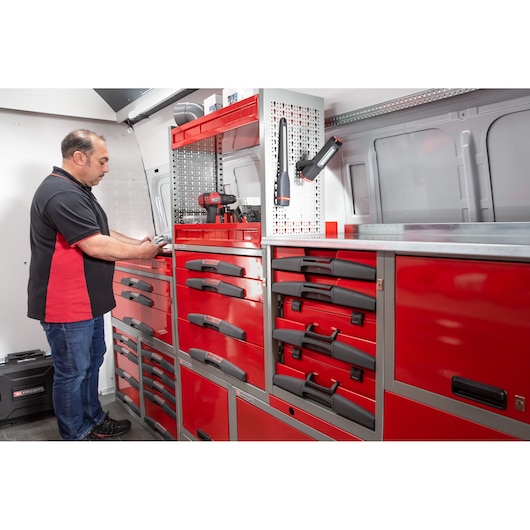 A person standing inside a well-organized work van equipped with multiple red Facom tool cabinets and drawers. Various tools and equipment are stored on the workbench and hung on a perforated panel.